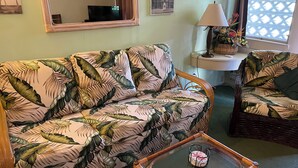 Ein Wohnzimmer mit einem sofa in floralem Muster, einem Korbstuhl, einem Glastisch, einer Lampe und einem an der Wand montierten Fernseher.