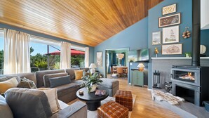 A modern living room with a fireplace, wooden ceiling, and a dining area visible through a glass door. - Spacious 4BD Beach House - Pet-Friendly - Hot Tub! (Manzanita)