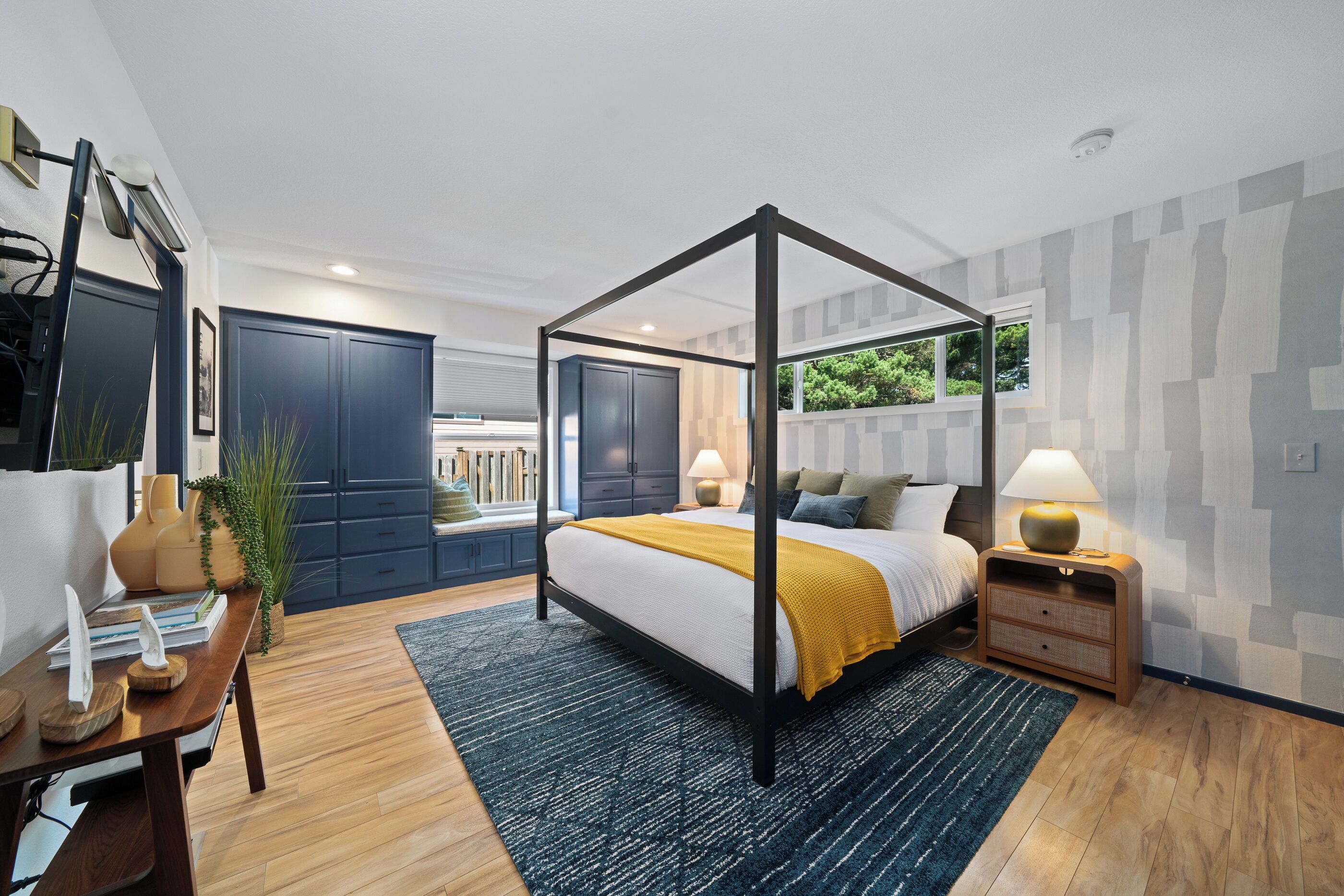 A modern bedroom with a four-poster bed, a desk with a chair, a nightstand, and a window with a view of greenery.