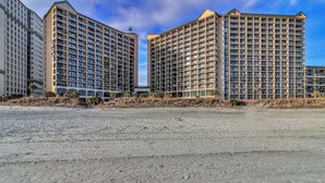 A beachfront hotel with multiple high-rise buildings, sandy beach in the foreground, and clear skies. - Oceanfront Condo just steps to the beach, Free parking and no resort fees! (North Myrtle Beach)