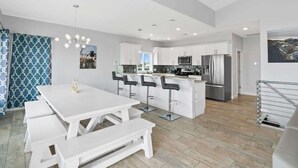 Una cocina moderna con armarios blancos, una isla central con taburetes y una zona de comedor con una mesa blanca y bancos.