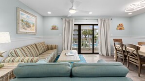 Un salon avec un canapé, une table à manger et des chaises, un ventilateur de plafond et une vue sur une piscine, visible à travers une porte vitrée.