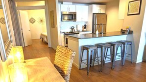 A modern kitchen with stainless steel appliances, a bar area with stools, and a dining area with a wooden table and a decorative chair. - Harbor Steps - 1 Blk to Pike Place Market, Waterfront - City Views! (Seattle)