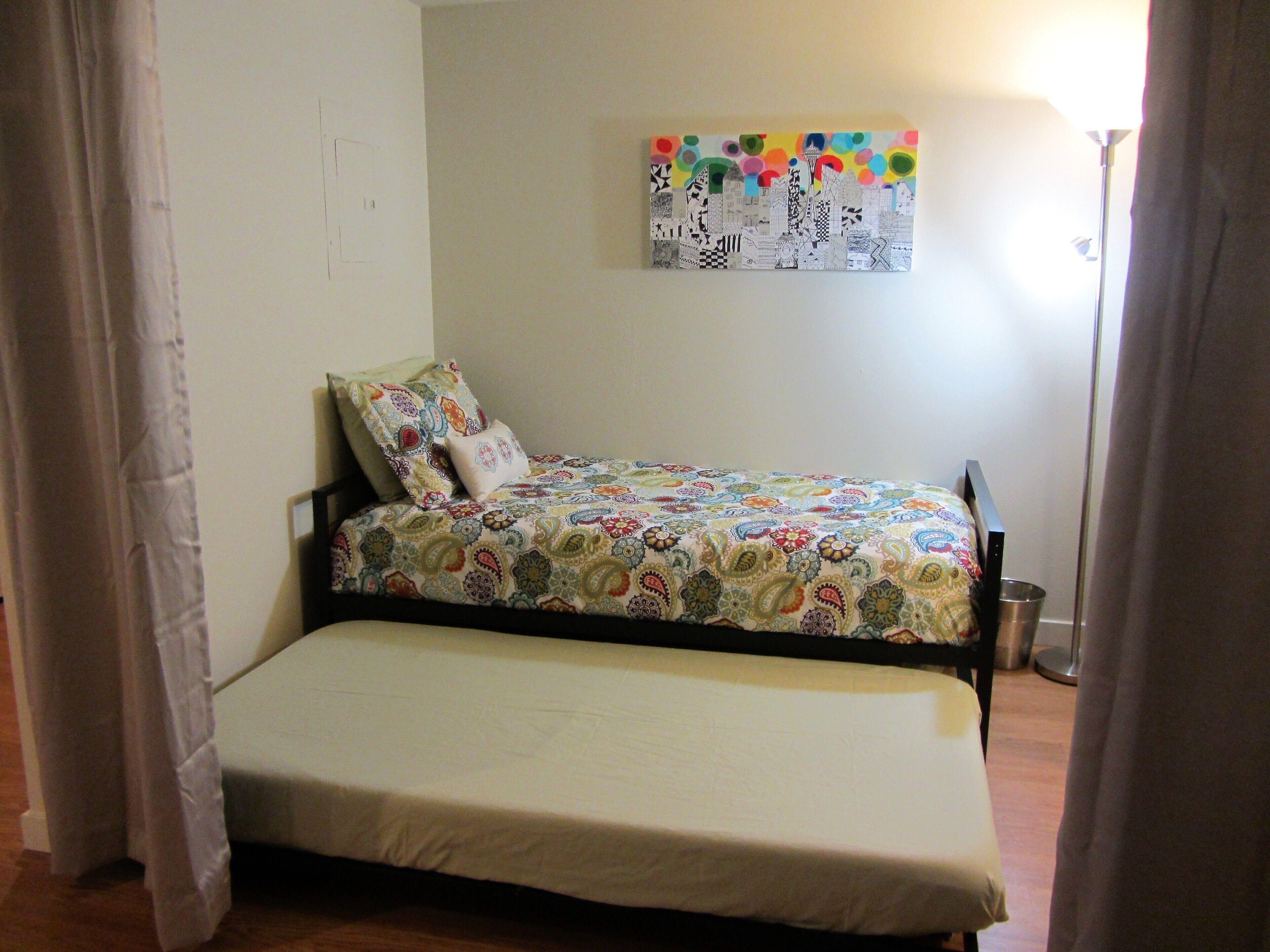 A small, single-bed room with a colorful quilt, a metal bucket, and a wall-mounted artwork.