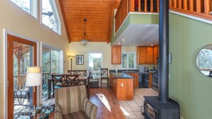 A spacious living area with a high ceiling, wooden flooring, a central seating area with a sofa and chairs, a glass coffee table, and a kitchen with wooden cabinets and a black wood stove. - Cozy Cottage Retreat,  Close to Yosemite, Beautiful Views, Hot Tub & More! (Mariposa)