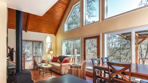 A spacious living area with a fireplace, large windows, and a dining area with wooden furniture. - Cozy Cottage Retreat,  Close to Yosemite, Beautiful Views, Hot Tub & More! (Mariposa)