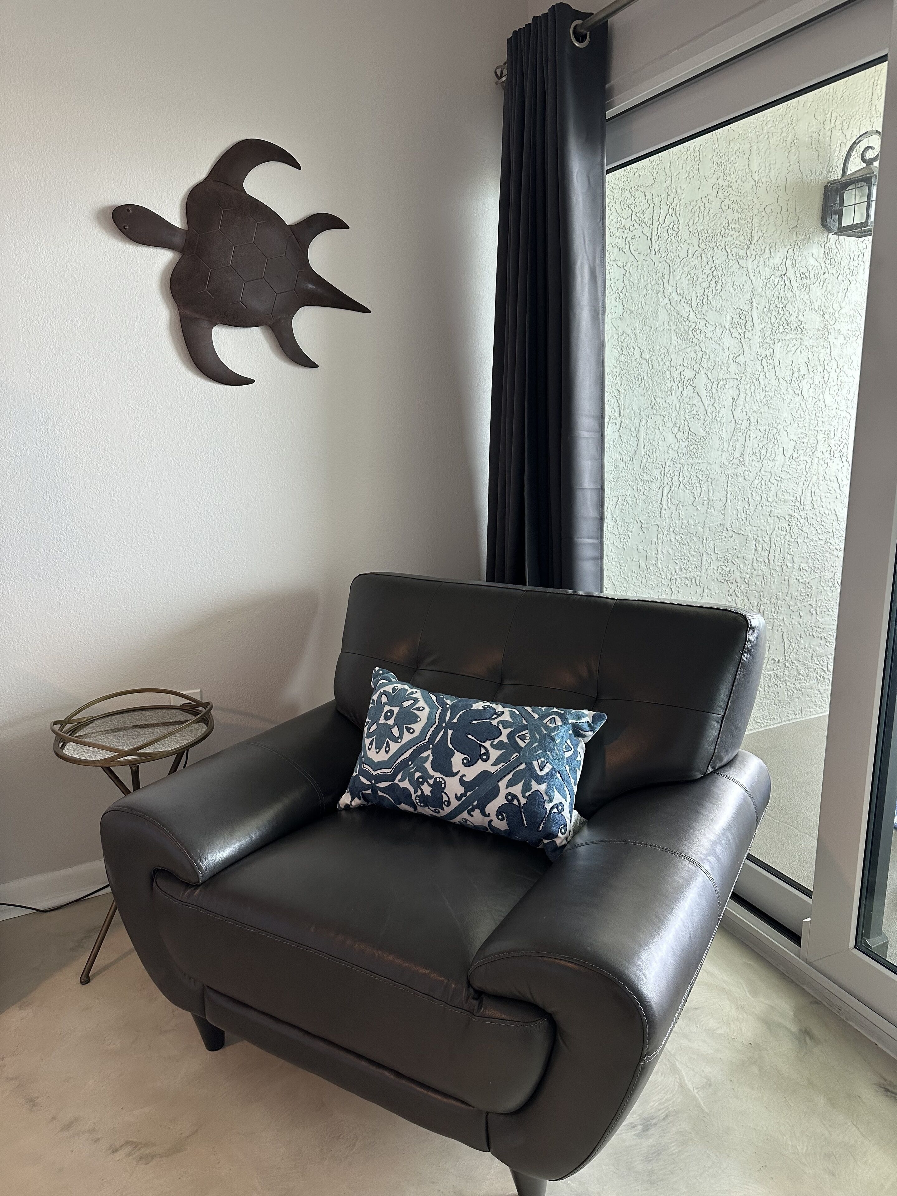 Un sillón de cuero negro con un cojín estampado, una mesita redonda pequeña y una decoración de tortuga montada en la pared en una habitación con un ventanal grande y pared con textura.