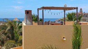 A rooftop terrace with a pergola, palm trees, and a view of the sea and historic buildings in the background. - Puesta Del Sol Top Unit 2BR Condo Private Rooftop+ Steps to Cerritos Surf Beach (Cerritos Beach)