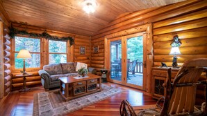Ein rustikales Holzhaus im Inneren, mit Holzboden, einer Couch, einem Couchtisch, einer Lampe und einem Essbereich, der durch eine Glastür sichtbar ist.