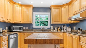 A kitchen with wooden cabinets, a stainless steel dishwasher, a granite countertop, and a window with a view of greenery. - Peek of Blue – 2nd Row Sea Pines Home, Pool & Spa, Sleeps 17, 800 Ft to Beach (Hilton Head Island)