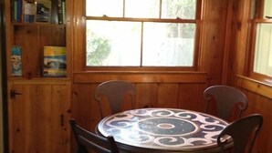 Un intérieur en bois avec une table ronde et quatre chaises, une bibliothèque et une fenêtre donnant sur un paysage verdoyant.