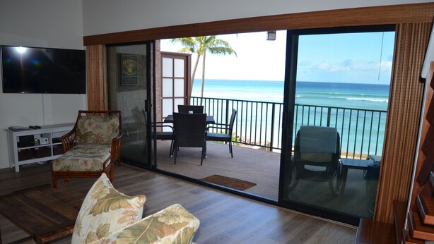 Een woonkamer met een grote televisie, een bank en een balkon met uitzicht op zee.