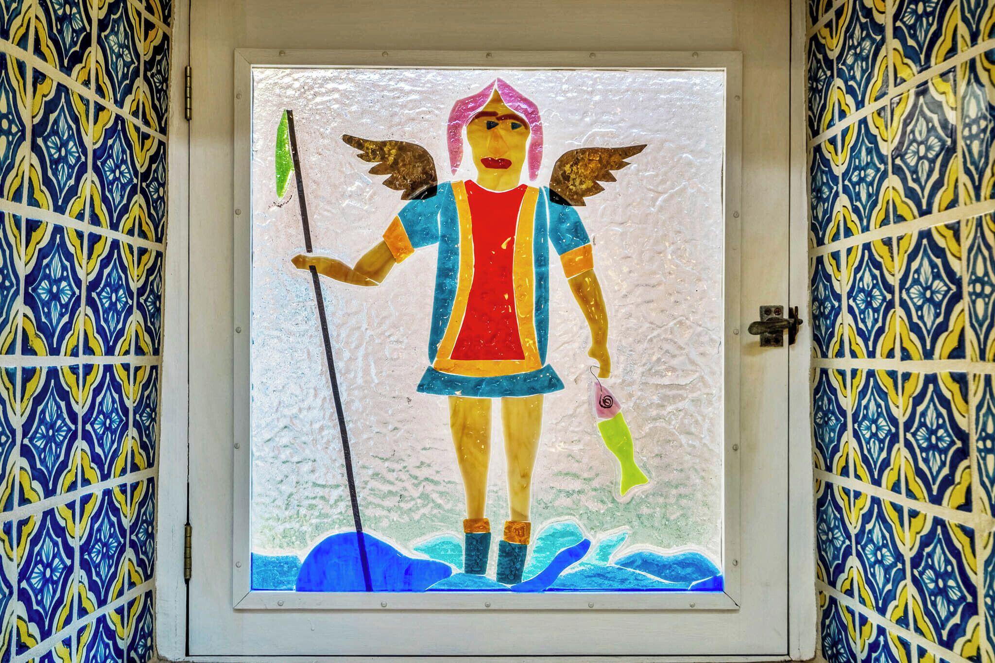 A stained glass window with a figure holding a staff and a fish, set against a patterned wall.