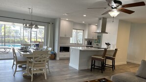 Una cocina moderna con armarios blancos, un comedor con mesa y sillas, un ventilador de techo y vistas al exterior.