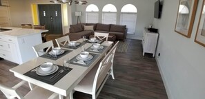 Ett modernt matområde med ett långt bord för fyra personer, vita stolar och en brun soffa i bakgrunden.
