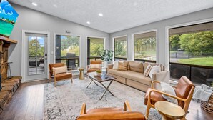 A spacious living area with a large sofa, two armchairs, and a coffee table. There are large windows offering views of a garden and a road.