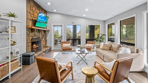 A spacious living room with a stone fireplace, comfortable seating, and large windows offering a view of the outdoors.