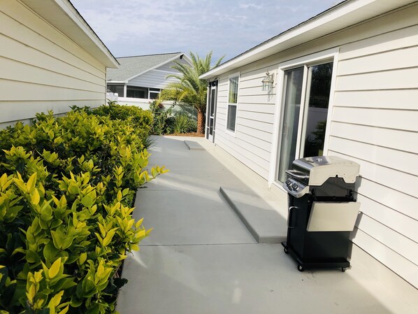 A narrow concrete pathway flanked by lush green bushes, leading to a white house with a sliding glass door and a barbecue grill on wheels.