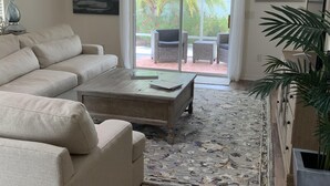 Un salon moderne avec un grand canapé d’angle, une table basse en bois et un tapis à motifs. Une porte-fenêtre coulissante donne accès à un patio extérieur équipé d’une chaise et d’une table.