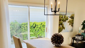 Un espace repas avec une table en bois, des chaises recouvertes d’un tissu beige, un lustre noir et une vue sur la verdure grâce à de grandes fenêtres.