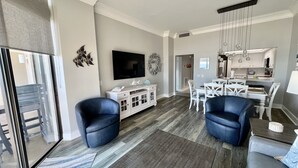 Un soggiorno moderno con una grande TV, due poltrone blu, un mobile bianco e un'area pranzo con sedie e tavolo bianchi.