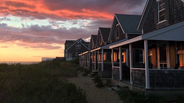 Una hilera de casas de playa con un sendero arenoso que las conecta, frente a un cielo al atardecer.
