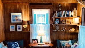 Interior acogedor de una cabaña con dos bancos de madera, una mesa central con una lámpara y una estantería llena de libros y objetos decorativos.