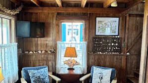 Una habitación acogedora con paredes y techo de madera, dos sillones azules con cojines decorativos, una mesita con una lámpara, una ventana con contraventanas azules y un televisor montado en la pared.