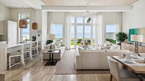 A modern living room with a large window overlooking a scenic view, a kitchen area with a bar, and a dining area with a table set for four. - On Beach Time |Lux Villa|Oceanfront|Heated Pool (Nassau)
