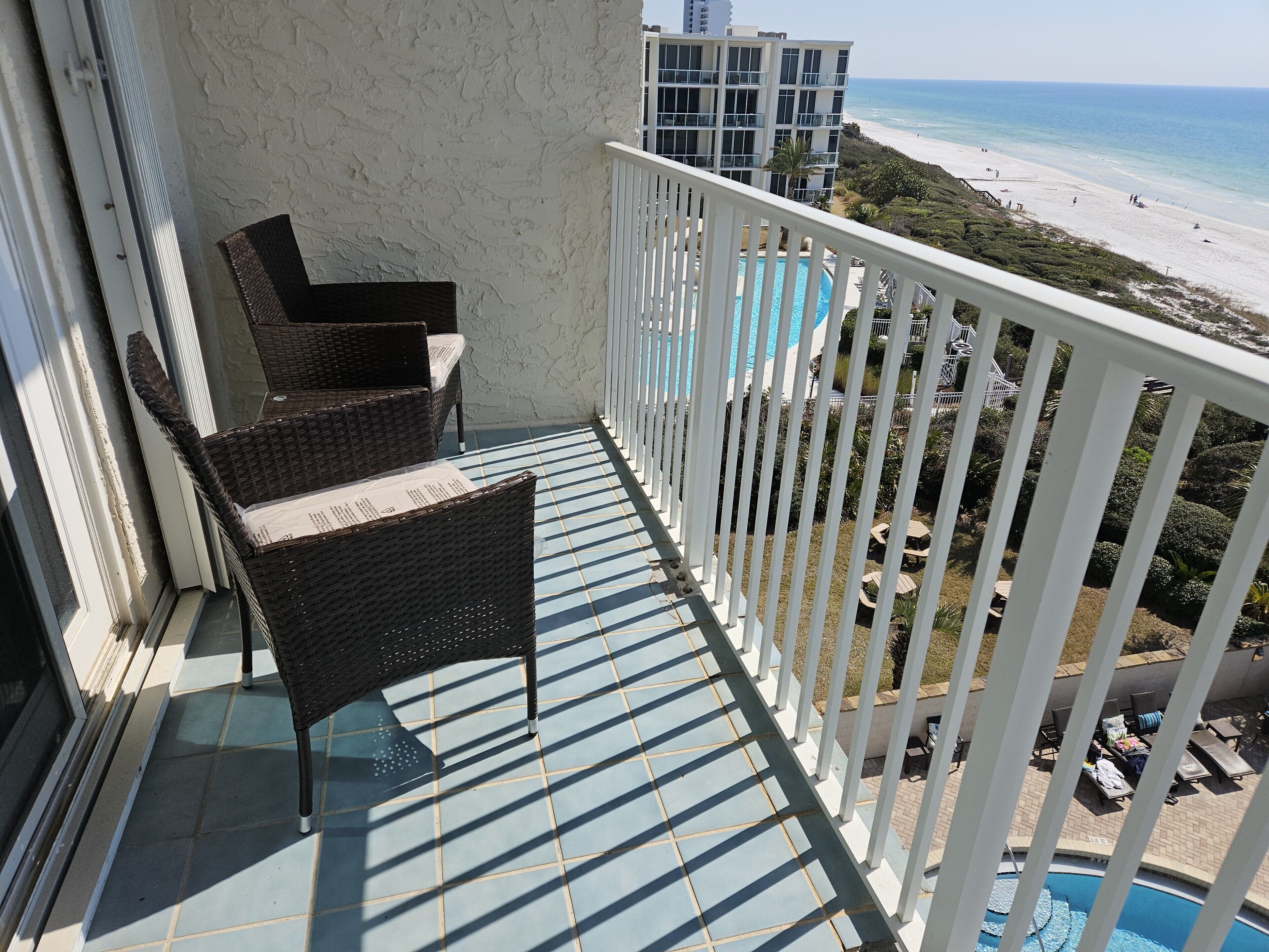 A balcony with wicker chairs, a pool, and a view of the beach and ocean.