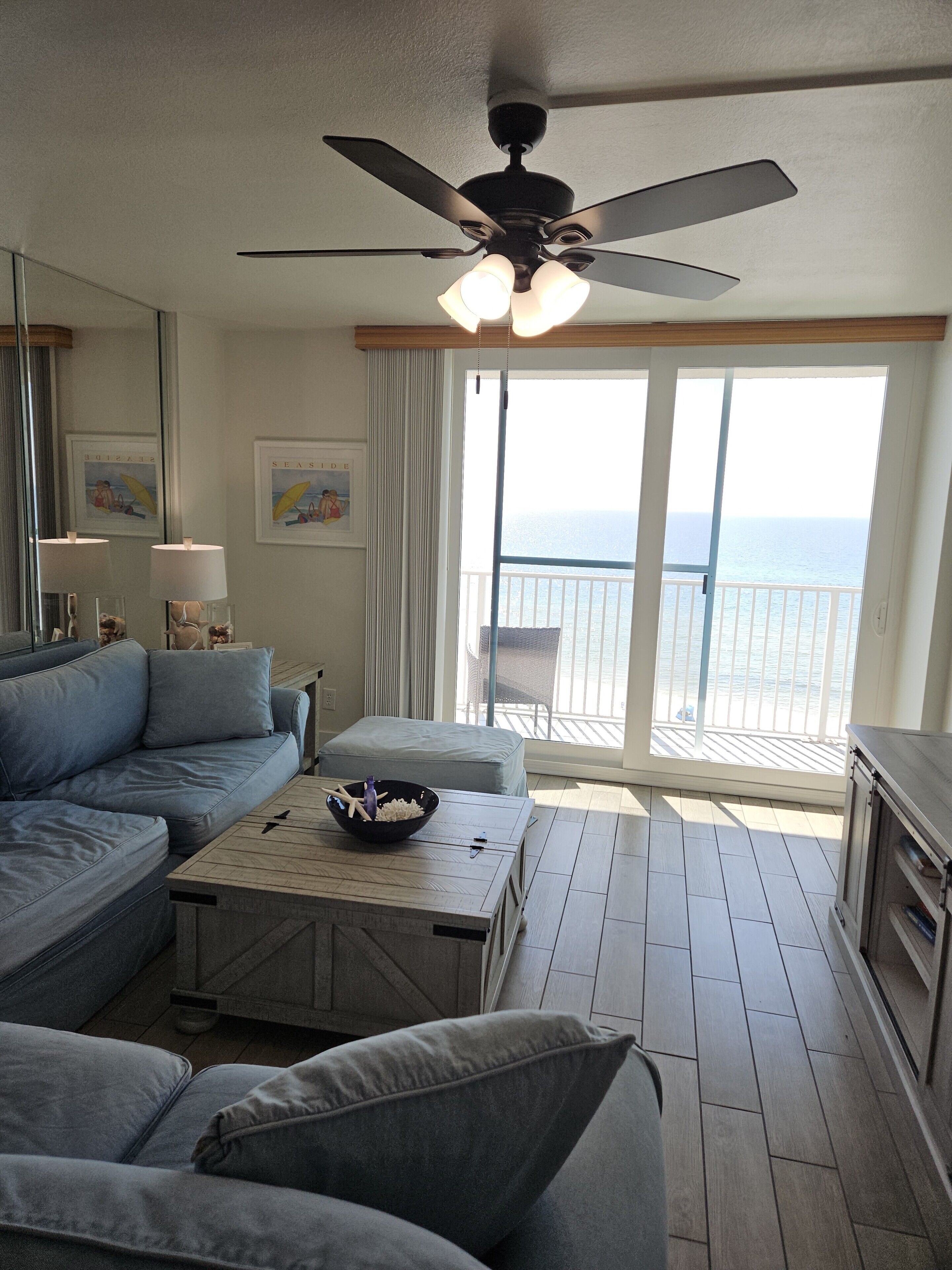 A modern living room with a sofa, a coffee table, and a ceiling fan. There is a balcony with a view of the ocean.