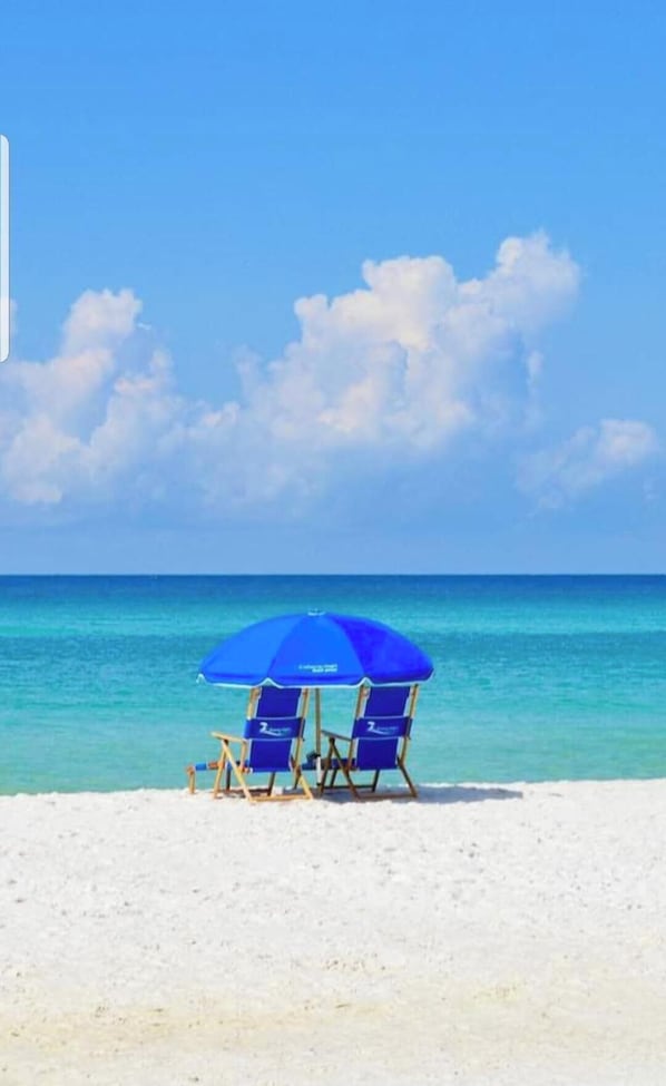 Une scène de plage avec un parasol bleu et deux chaises de plage sur le sable.