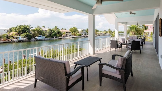 A covered outdoor area with wicker furniture, a table, and a view of a body of water and distant buildings. - Grouper House: 4 Masters/6 BA, Pool/Dock, BoatRamp (Marathon)