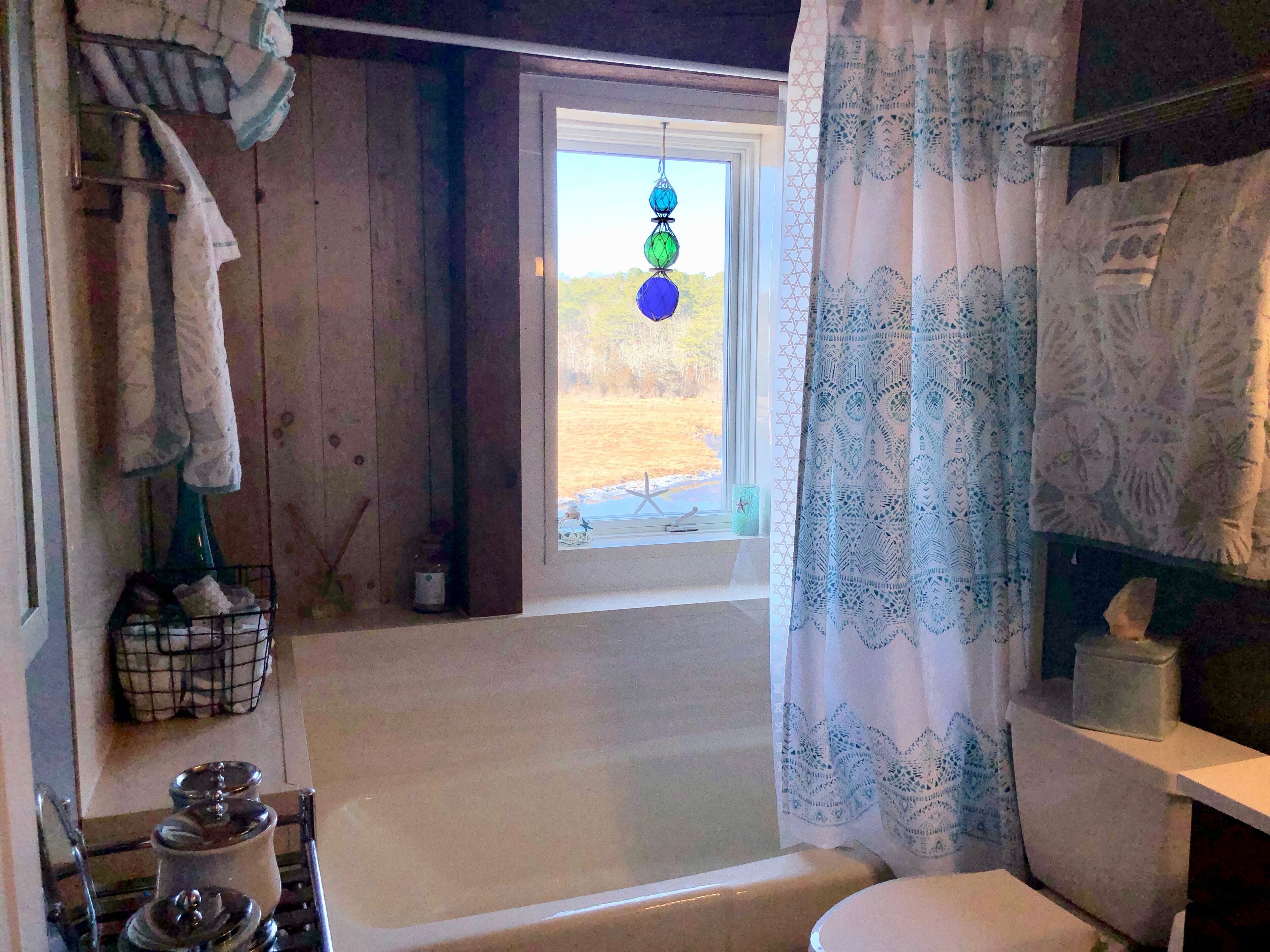 A bathroom with a bathtub, a window with a view of a grassy field, and a curtain with a geometric pattern.
