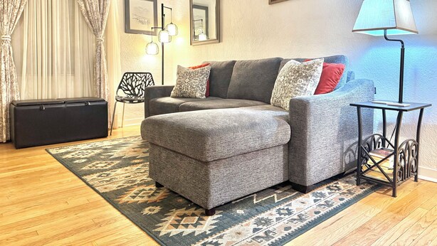 A living room with a grey sofa, patterned cushions, a patterned rug, a black side table with a lamp, and a framed picture on the wall. - Perfect family friendly home in Nob Hill (Albuquerque)