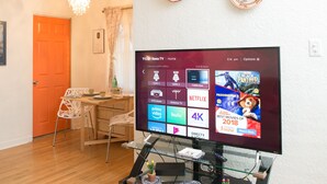 A living room with a flat-screen TV displaying a Roku TV interface, a dining area with a wooden table and chairs, and a bright orange door. - Perfect family friendly home in Nob Hill (Albuquerque)