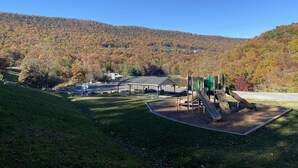Ein Spielplatz mit Rutschen und Klettergerüsten, umgeben von einer Rasenfläche, mit herbstlich gefärbten Bäumen im Hintergrund.