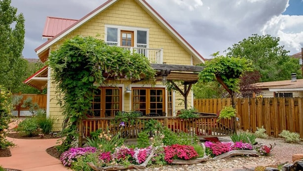 Ett tvåvåningshus med en veranda, omgivet av en trädgård med olika växter och blommor.