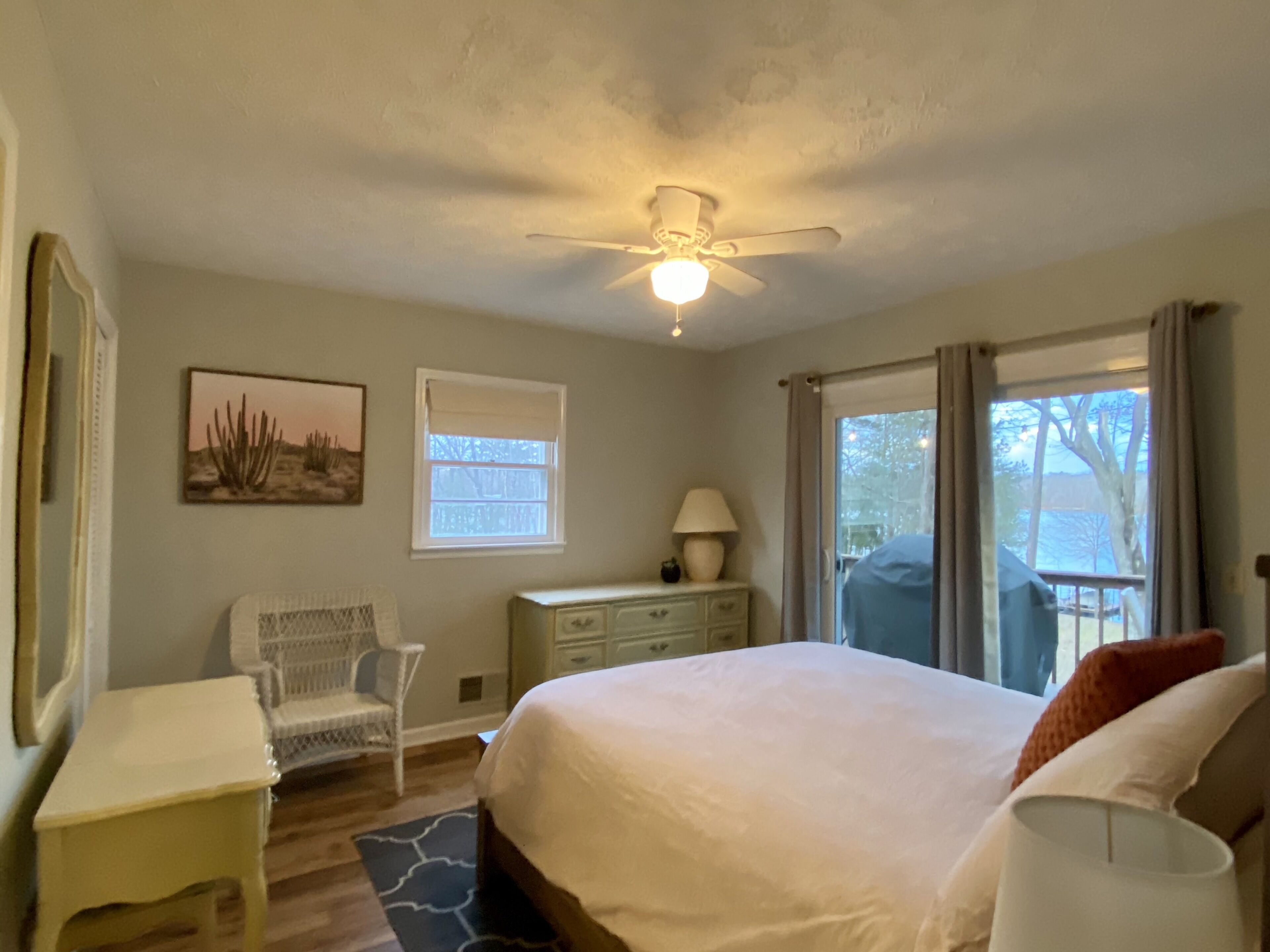 Une chambre à coucher comprenant un lit, une chaise, un bureau, une lampe, un ventilateur de plafond, une fenêtre et une vue sur l’extérieur.
