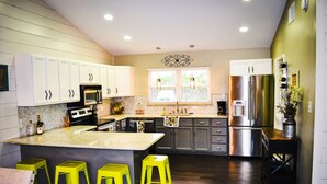 A modern kitchen with white cabinets, a central island with bar stools, stainless steel appliances, and a window with blinds. - 7-Acre Hill Country Haven w/ POOL-Guadalupe River (Bergheim)