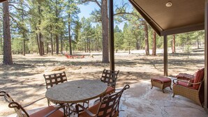 A covered outdoor seating area with a round table and wicker chairs, surrounded by a forested landscape.