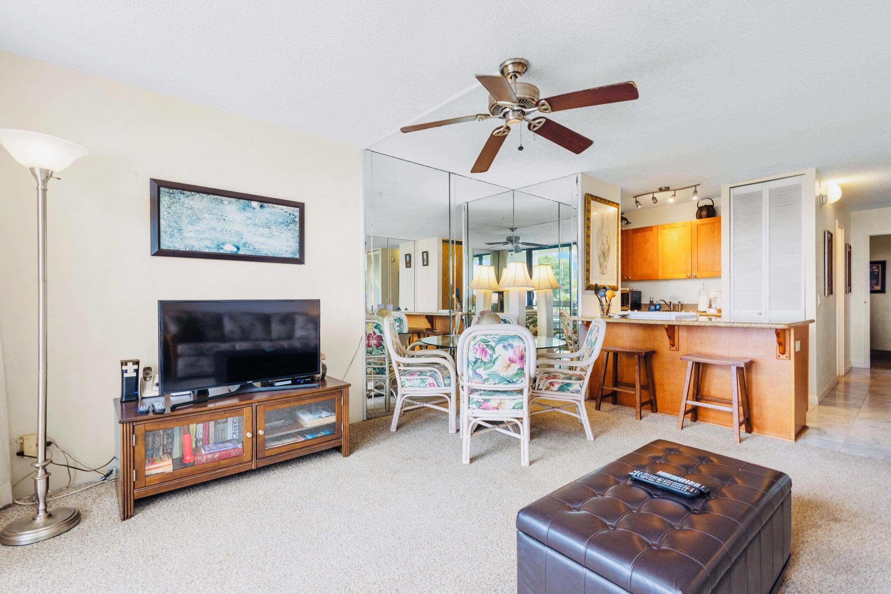 A living room with a television, a ceiling fan, a dining area with chairs, and a kitchen area with a bar.