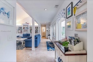 Una sala de estar moderna con un sofá azul, una mesa de centro y una estantería. Hay un comedor con una mesa y sillas de madera. La habitación tiene un ventanal grande y un ventilador de techo.