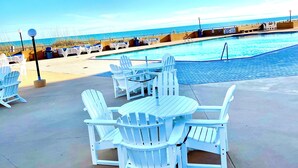 A poolside area with white Adirondack chairs and a table, overlooking a clear blue pool and the ocean.