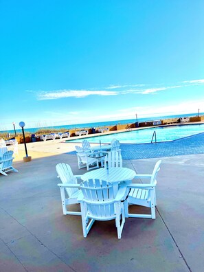 A poolside area with white Adirondack chairs and a table, overlooking a clear blue pool and the ocean.