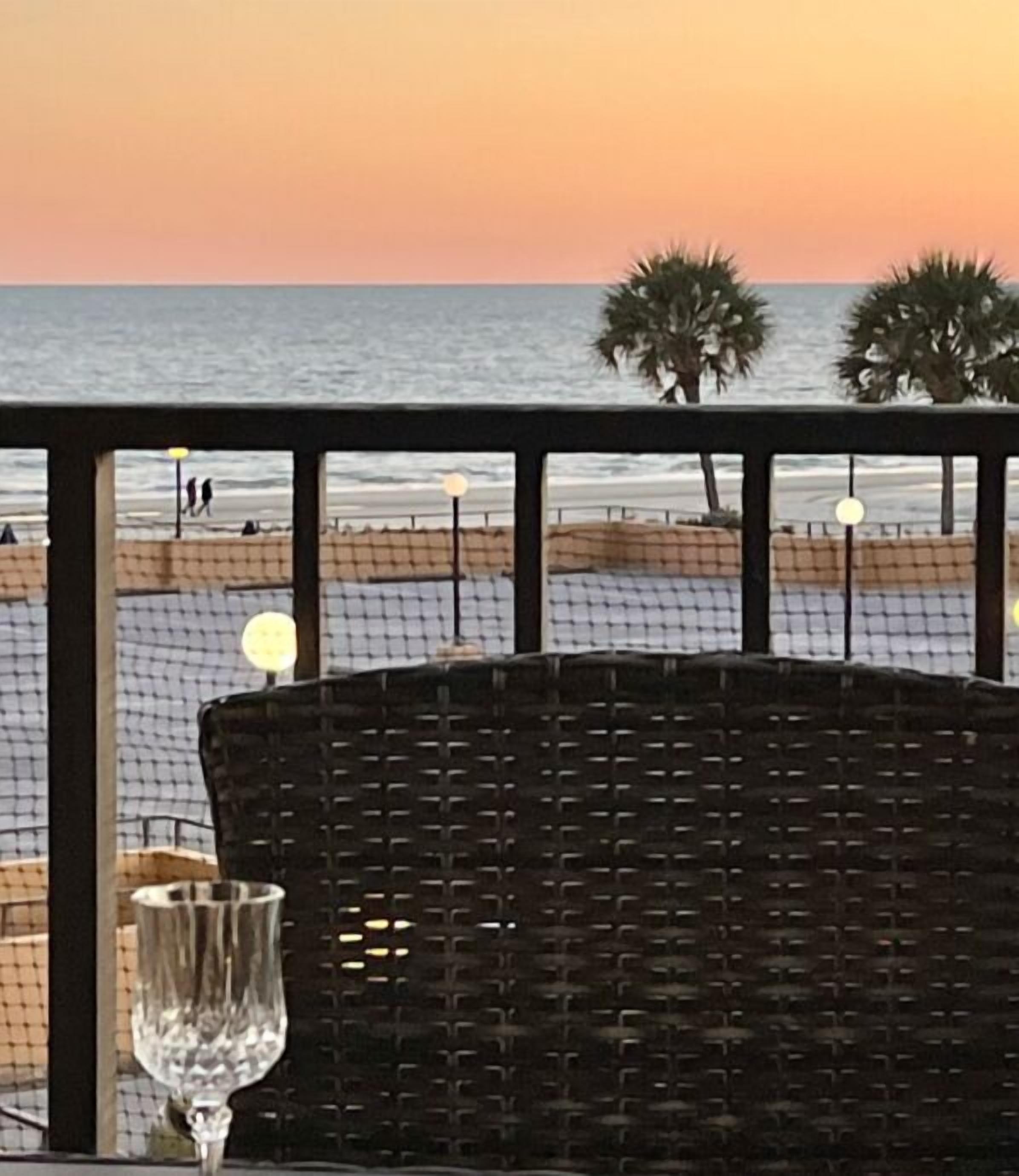 Een rieten stoel met een glas op een balkon met uitzicht op een strand bij zonsondergang.
