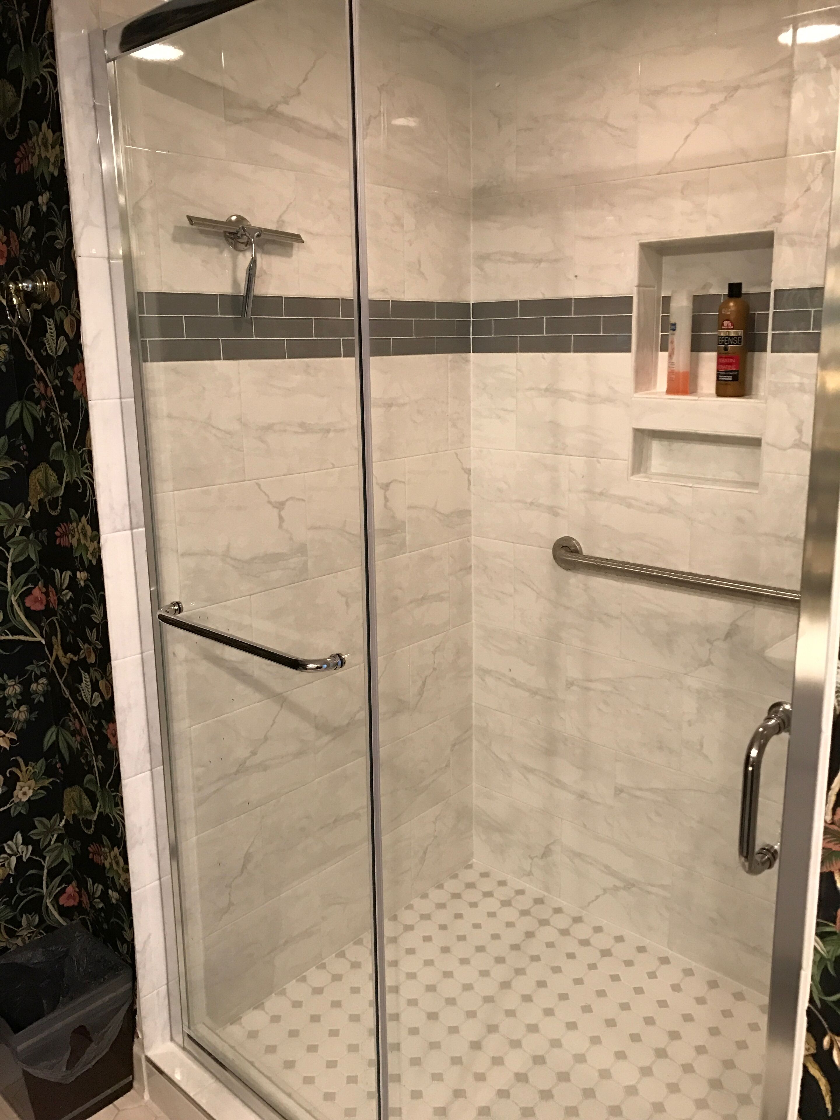 A glass-enclosed shower with a tiled floor, a handrail, and a shelf with toiletries.