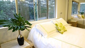 A bedroom with a large window, a bed with white bedding, a green pillow, a potted plant, and a wicker side table. - Gorgeous! The Waterfall Retreat At Reed's Island (Hilo)