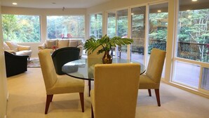 A spacious living room with a glass table surrounded by chairs, large windows offering a view of the outdoors, and a potted plant on the table. - Gorgeous! The Waterfall Retreat At Reed's Island (Hilo)