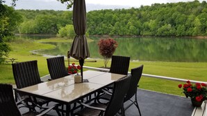 Een terras met tafel en stoelen met uitzicht op een meer, omgeven door groen.